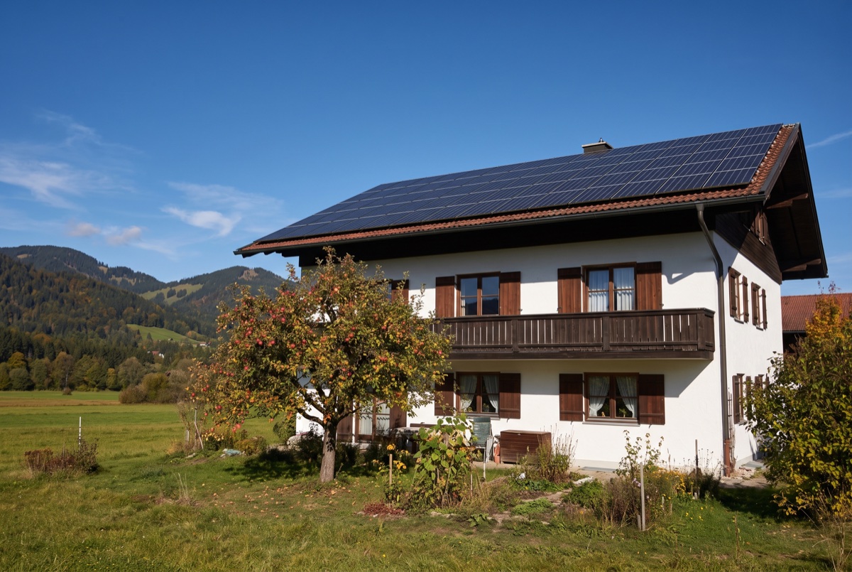 Weißes Einfamilienhaus mit Holzbalkon in Oberbayern, das Süddach voll mit Photovoltaikmodulen belegt, dahinter die Alpen